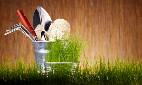 The image displays a close-up view of a garden scene with two small garden buckets placed on a lush, green grassy surface. One bucket contains a collection of gardening tools, including a trowel, a hand rake, and a pair of pruning shears, all with metal blades and red or black handles. The second bucket holds a bunch of bright green grass or small seedlings, along with natural twine or string coiled nearby. The background features a warm-toned wooden fence or deck, providing a textured backdrop that contrasts with the vibrant green of the grass in the foreground. The scene suggests preparation for gardening or lawn maintenance, aligning with landscape and outdoor upkeep services, and the natural light indicates a clear, sunny day in an outdoor garden space likely located in or around Hanwell, London. This detailed and natural depiction of gardening tools and fresh grass supports the gardening services offered by Hedge Trimming Hanwell, emphasizing outdoor care and lawn management.
