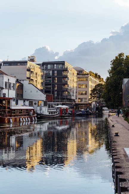 A tranquil scene along the Grand Union Canal in Hanwell, showing calm water reflecting the modern residential buildings and a partly cloudy sky. The canalbank features a paved walkway with a row of bo