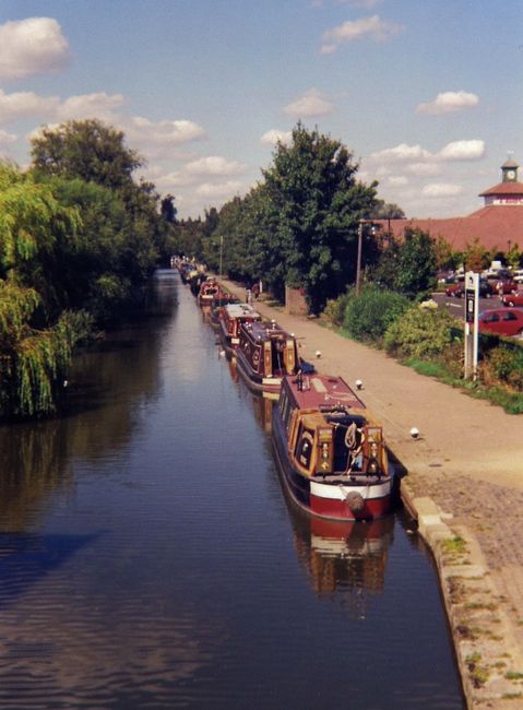 A tranquil scene along the Grand Union Canal in Hanwell, showing calm water reflecting the modern residential buildings and a partly cloudy sky. The canalbank features a paved walkway with a row of bo